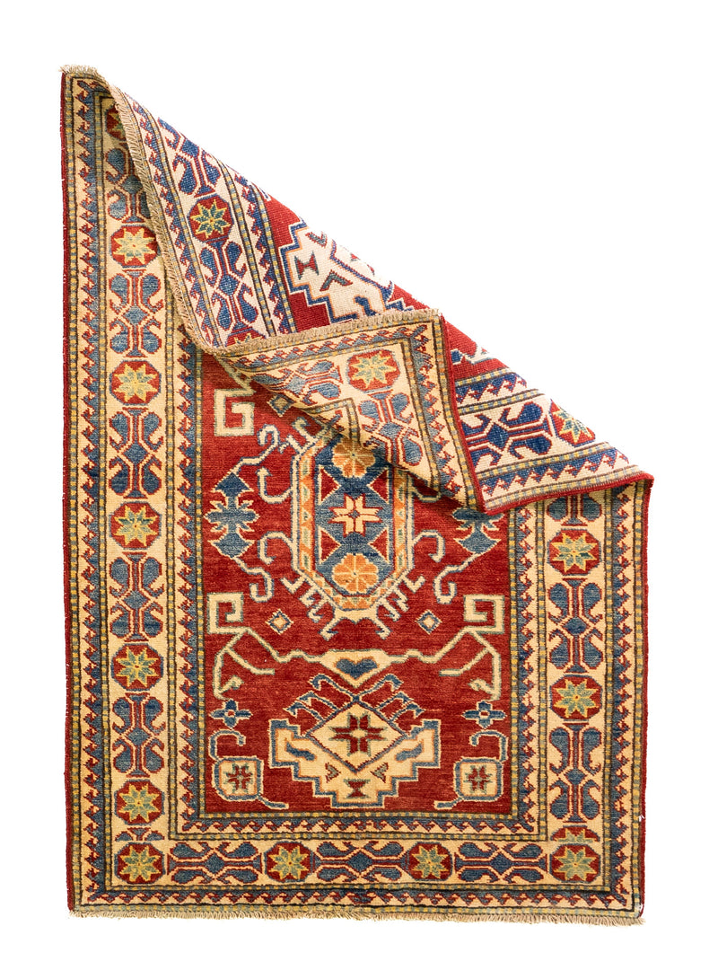 Alfombra Caucasica Kazak ladrillo y beige de 1.09 x 1.58 mts, vista completa con una esquina doblada; el diseño muestra medallones geométricos escalonados sobre un campo ladrillo con guardas claras y pequeños acentos