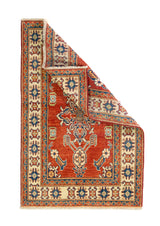 Alfombra Caucasica Kazak ladrillo y beige de 0.94 x 1.45 mts, vista completa con una esquina doblada; el diseño muestra un motivo central estilizado sobre un campo ladrillo enmarcado por guardas beige
