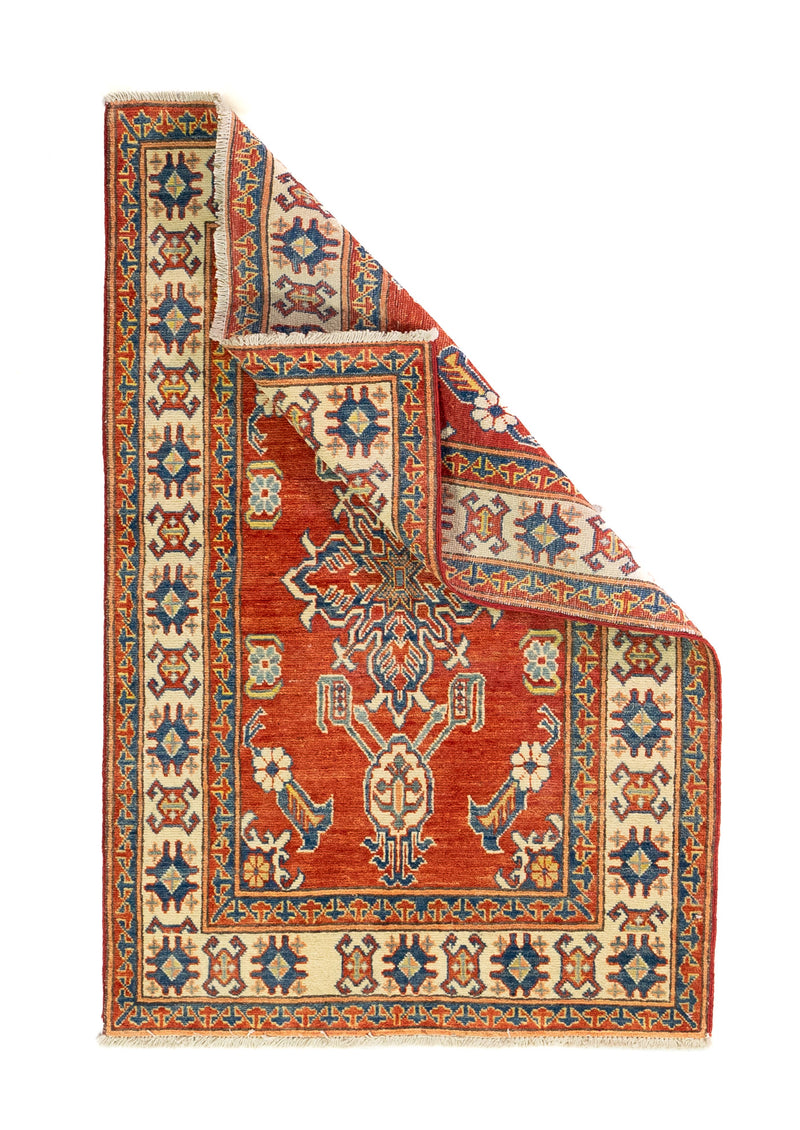 Alfombra Caucasica Kazak ladrillo y beige de 0.94 x 1.45 mts, vista completa con una esquina doblada; el diseño muestra un motivo central estilizado sobre un campo ladrillo enmarcado por guardas beige