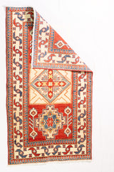 Alfombra Caucasica Kazak rojo y beige de 0.91 x 1.60 mts, vista completa con una esquina doblada; el diseño muestra tres medallones geométricos destacados sobre un campo rojo con amplias guardas beige