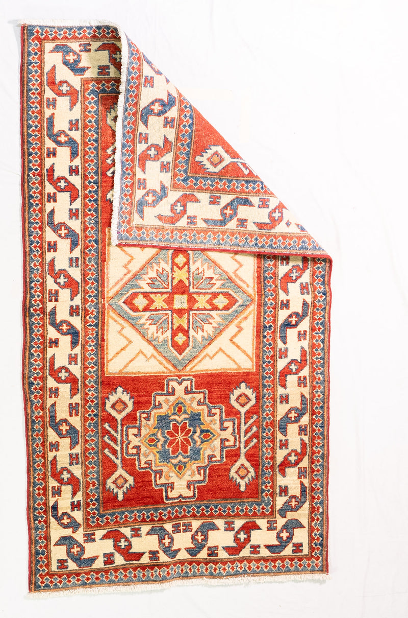Alfombra Caucasica Kazak rojo y beige de 0.91 x 1.60 mts, vista completa con una esquina doblada; el diseño muestra tres medallones geométricos destacados sobre un campo rojo con amplias guardas beige