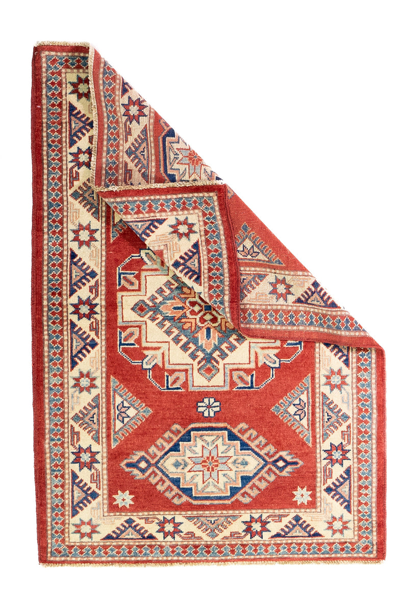 Alfombra Caucasica Kazak ladrillo y beige de 1.04 x 1.47 mts, vista completa con una esquina doblada; el diseño muestra tres medallones geométricos principales sobre un campo ladrillo con borde beige
