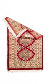 Alfombra Persa Yamouth beige y rojo de 0.61 x 1.09 mts, vista completa con una esquina doblada; el diseño muestra un motivo enlazado rojo de gran escala sobre un campo beige y un borde rojo intenso