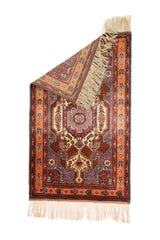 Alfombra Caucasica Kazak marrón y naranja de 0.97 x 1.63 mts, vista completa con una esquina doblada; el diseño muestra un medallón central y motivos angulares sobre un campo marrón con guardas naranjas