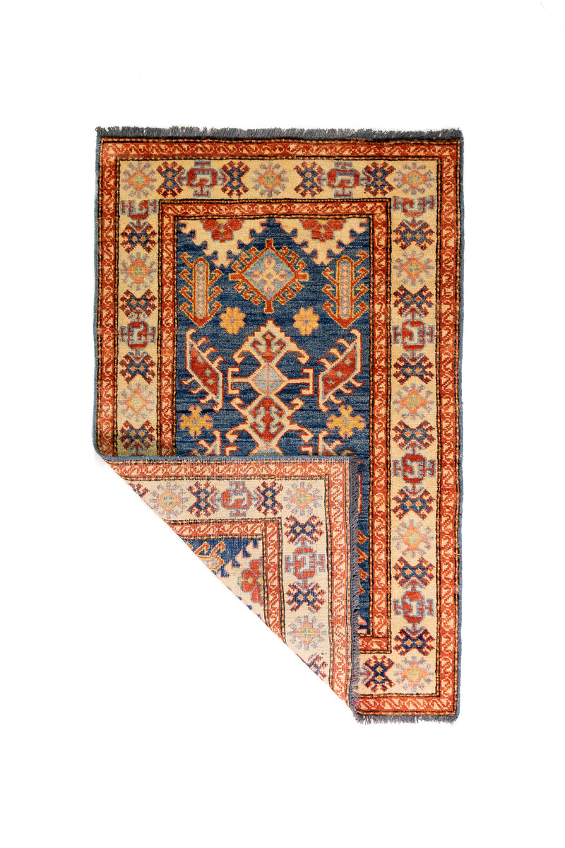 Alfombra Caucasica Kazak beige y azul de 0.83 x 1.30 mts, vista completa con una esquina doblada; el diseño muestra medallones geométricos azules sobre un campo beige con guardas de acento ladrillo