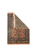 Alfombra Persa Bukara rosa viejo y gris de 0.63 x 0.90 mts, vista con una esquina doblada para mostrar guarda, reverso y estructura visual