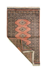Alfombra Persa Bukara rosado y gris de 0.94 x 1.60 mts, vista con una esquina doblada para mostrar guarda, reverso y estructura visual
