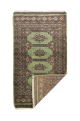 Alfombra Persa Bukara verde y rosa de 0.76 x 1.30 mts, vista con una esquina doblada para mostrar guarda, reverso y estructura visual