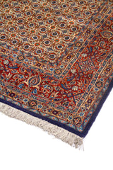 Detalle en perspectiva de alfombra con pequeño patrón geométrico repetido, borde rojo ornamental y terminación azul oscuro.