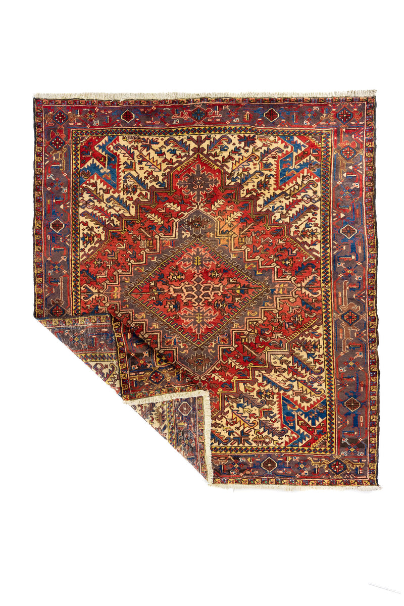 Alfombra geométrica en rojo y beige con borde azul; esquina doblada para mostrar el grosor, tejido y flecos.