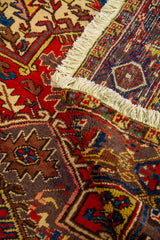 Detalle con la alfombra doblada mostrando reverso, costura del borde y densidad del tejido en tonos rojo, azul y beige.