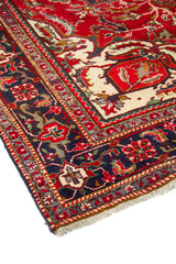 Vista en ángulo de alfombra oriental con campo rojo, elementos decorativos marfil y borde geométrico en azul y rojo.