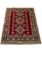 Alfombra Caucasica Kazak rojo y beige de 1.17 x 1.47 mts, vista en perspectiva del campo y las guardas; el diseño muestra medallones romboidales apilados sobre un campo rojo con amplias guardas beige