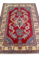 Alfombra Caucasica Kazak bordó y beige de 1.14 x 1.65 mts, vista en perspectiva del campo y las guardas; el diseño muestra un gran motivo central de lectura geométrica sobre un campo bordó y guardas beige