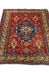 Alfombra Caucasica Kazak ladrillo y beige de 1.07 x 1.45 mts, vista en perspectiva del campo y las guardas; el diseño muestra un medallón central azul rodeado por motivos geométricos sobre un campo ladrillo