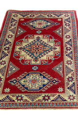 Alfombra Caucasica Kazak ladrillo y beige de 1.04 x 1.47 mts, vista en perspectiva del campo y las guardas; el diseño muestra tres medallones geométricos principales sobre un campo ladrillo con borde beige