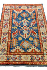 Alfombra Caucasica Kazak beige y azul de 0.83 x 1.30 mts, vista en perspectiva del campo y las guardas; el diseño muestra medallones geométricos azules sobre un campo beige con guardas de acento ladrillo