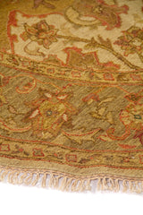 Detalle de alfombra redonda con guarda ornamental, motivos florales y flecos en tono beige.
