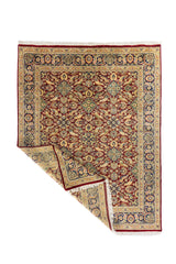 Alfombra persa roja con diseño floral y esquina doblada, mostrando el reverso y detalle de los flecos blancos.