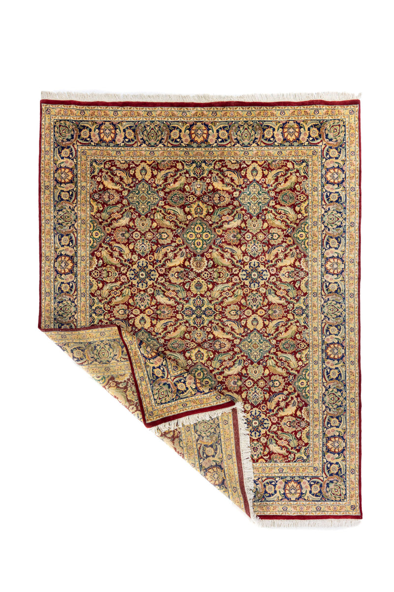 Alfombra persa roja con diseño floral y esquina doblada, mostrando el reverso y detalle de los flecos blancos.