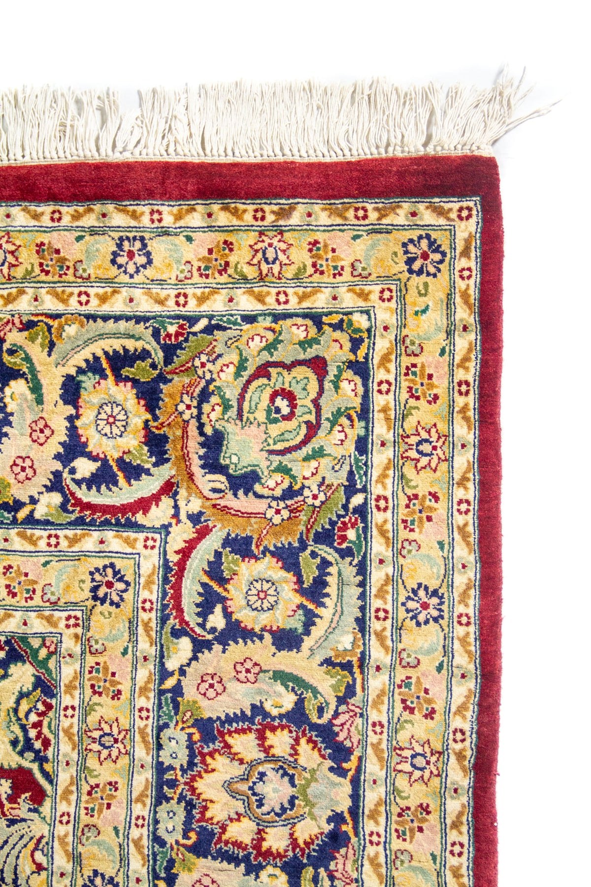 Detalle de esquina de alfombra oriental con borde rojo, guarda azul y beige, motivos florales y flecos blancos.
