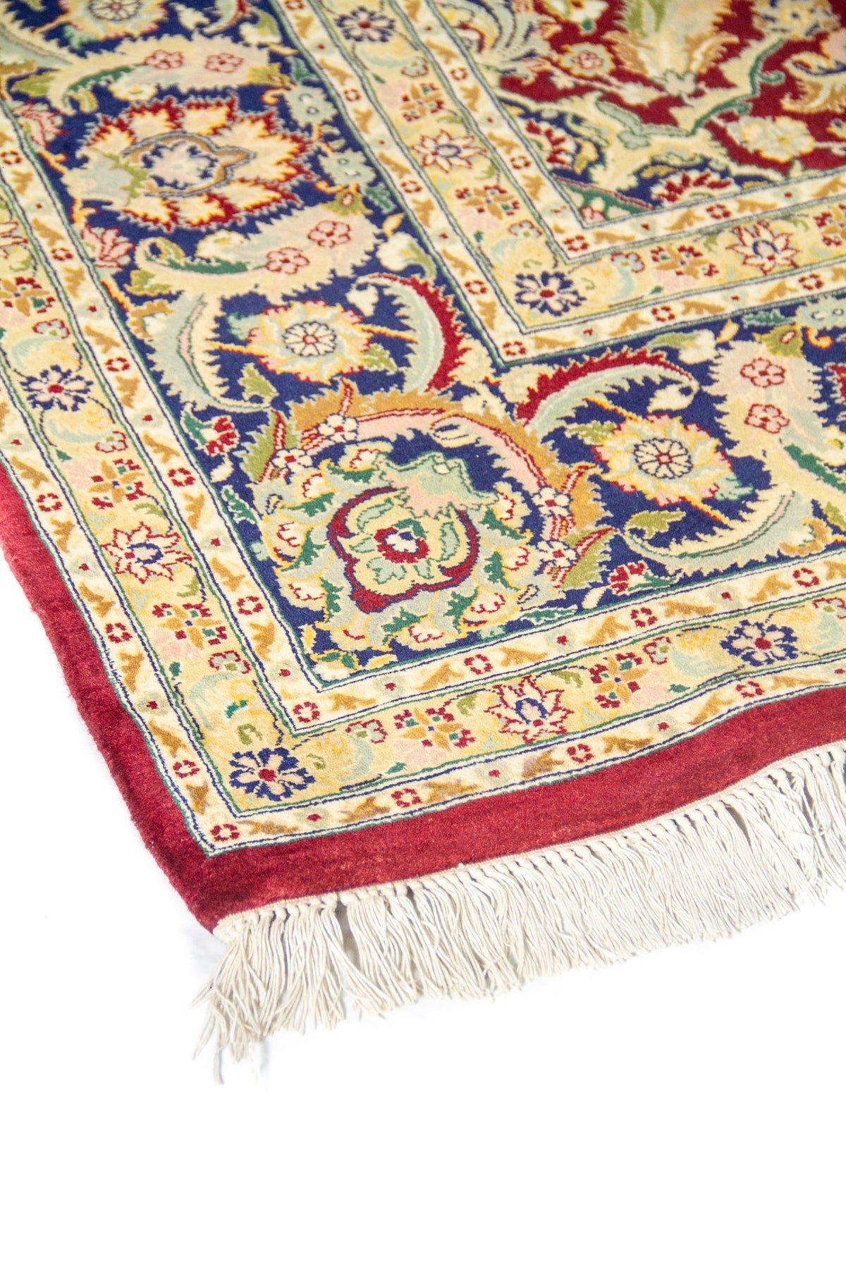 Vista en ángulo de alfombra clásica roja con patrón floral, guarda azul marino y flecos en uno de los extremos.