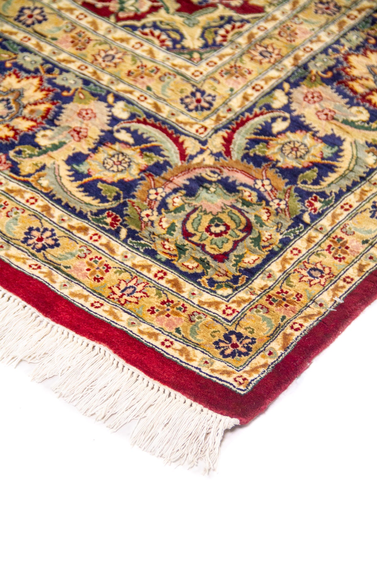 Primer plano de esquina de alfombra persa con fondo rojo, ornamentos florales en azul, beige y verde, y terminación con flecos.