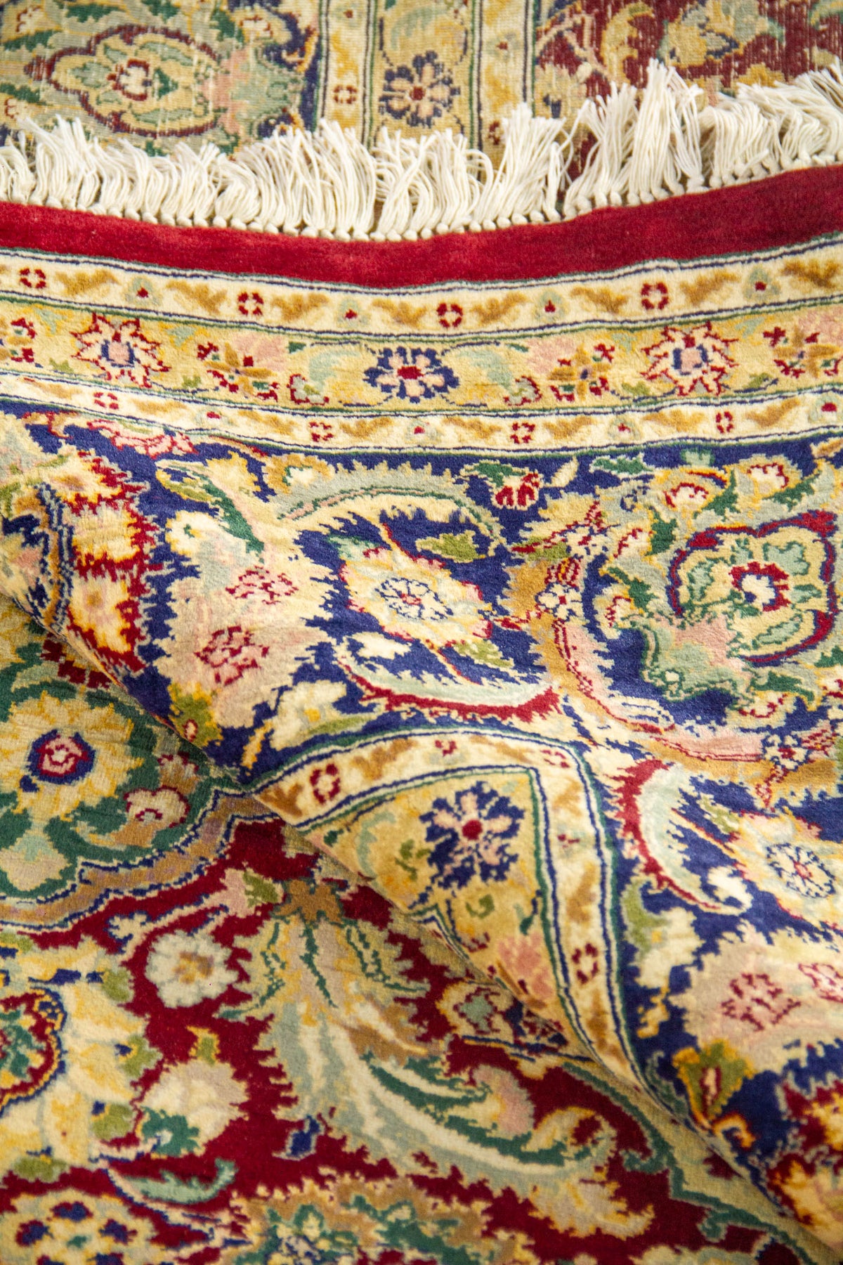 Detalle de alfombra doblada mostrando textura, diseño floral en rojo y azul, guardas ornamentales y flecos blancos.
