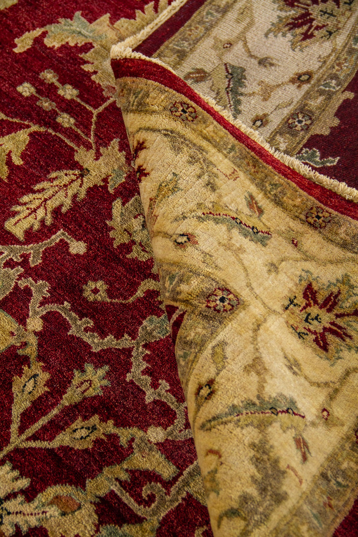 Alfombra doblada mostrando la textura del tejido, el contraste entre el fondo rojo y la guarda beige, y sus flecos.