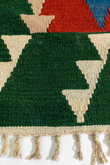 Primer plano del borde inferior izquierdo de la alfombra kilim con figuras triangulares color beige sobre fondo verde.