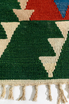 Primer plano del borde inferior izquierdo de la alfombra kilim con figuras triangulares color beige sobre fondo verde.