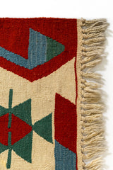 Detalle de la esquina superior derecha con patrón triangular verde, azul y rojo sobre fondo beige, acompañado de flecos tejidos a mano.
