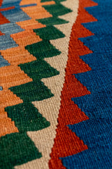 Detalle cerrado del tejido de la alfombra kilim mostrando la textura de los motivos geométricos verdes y naranjas sobre fondo beige y azul.
