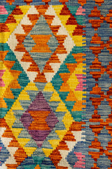 Primer plano del centro de la alfombra kilim con rombos entrelazados en amarillo, morado, azul y naranja, rodeados de patrones escalonados.