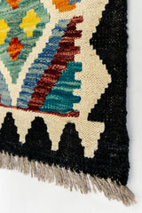 Primer plano del borde de la alfombra kilim con diseño en zigzag negro y remate de flecos grises.