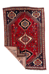 Alfombra roja de estilo Kazak con una esquina doblada mostrando el reverso tejido, el grosor y el acabado del borde.