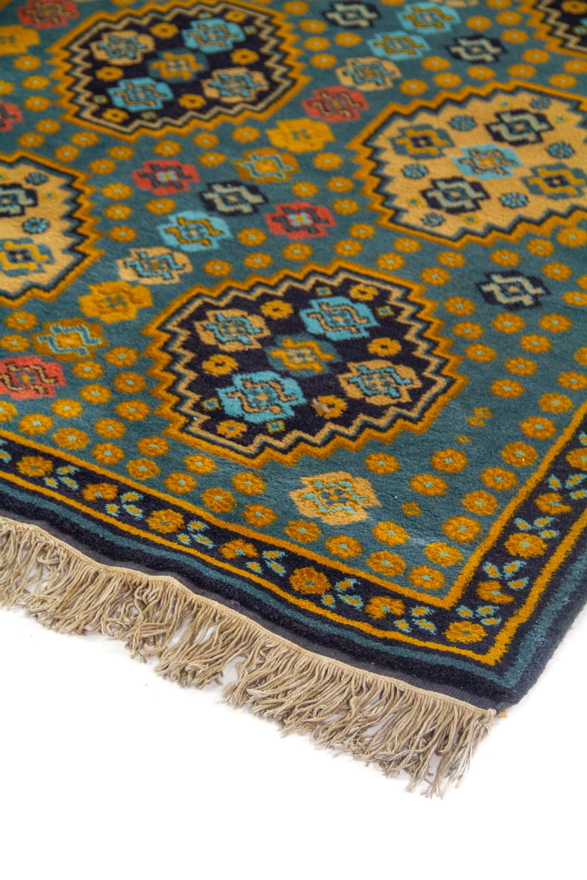 Detalle en perspectiva de alfombra con fondo azul suave, figuras geométricas repetidas y borde decorativo en azul oscuro con acentos dorado.