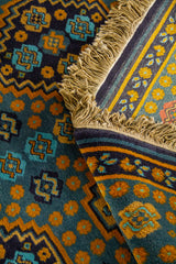 Detalle de alfombra doblada mostrando la textura del tejido, el reverso, los flecos y los contrastes entre azul, beige, dorado y celeste.