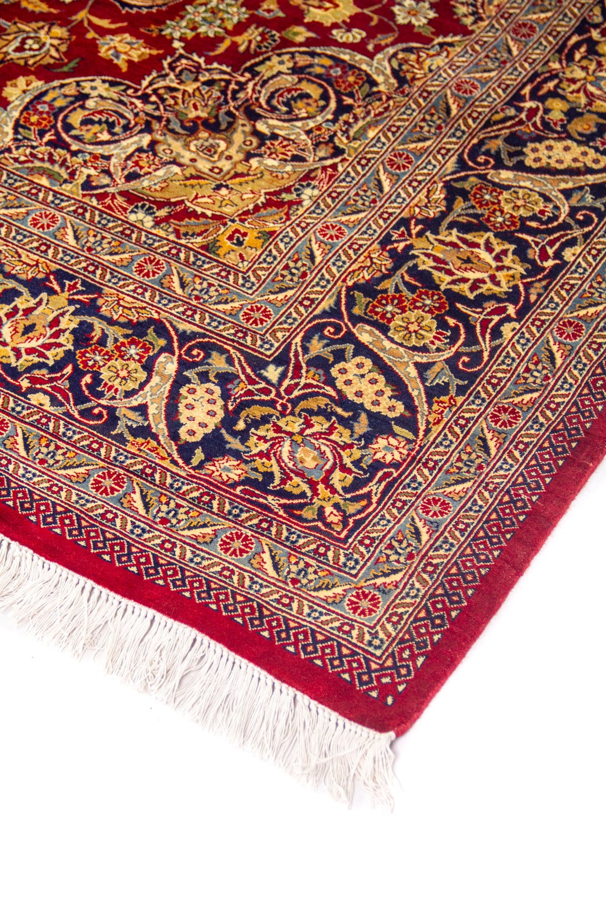 Vista en ángulo de la esquina de alfombra roja con borde azul decorado, patrón floral ornamental y acabado con flecos.