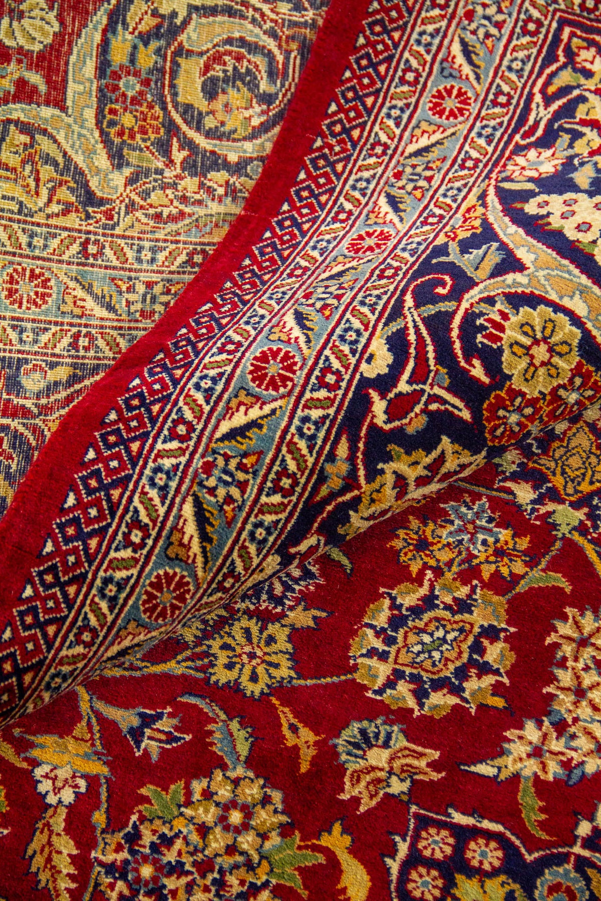 Primer plano de alfombra doblada mostrando textura, reverso del tejido y motivos florales en rojo, azul y beige.