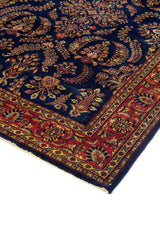 Detalle en perspectiva de alfombra azul marino con borde rojo, diseño floral y terminación con flecos beige.