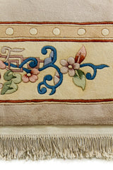Detalle del borde floral de la alfombra con flores rosadas, hojas verdes y trazos azul profundo sobre fondo beige.