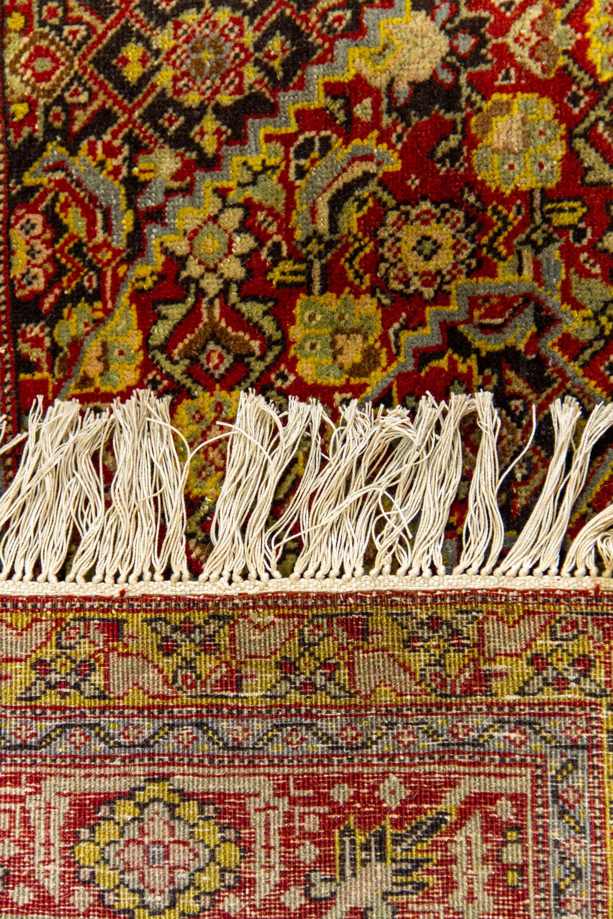 Detalle cercano de los flecos y el borde inferior de una alfombra oriental, con motivos florales sobre fondo rojo intenso y amarillo mostaza.