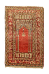 Vista superior de una alfombra de oración con campo rojo liso, marco decorativo y diseño arquitectónico inspirado en un mihrab, en tonos beige, marrón y oliva.