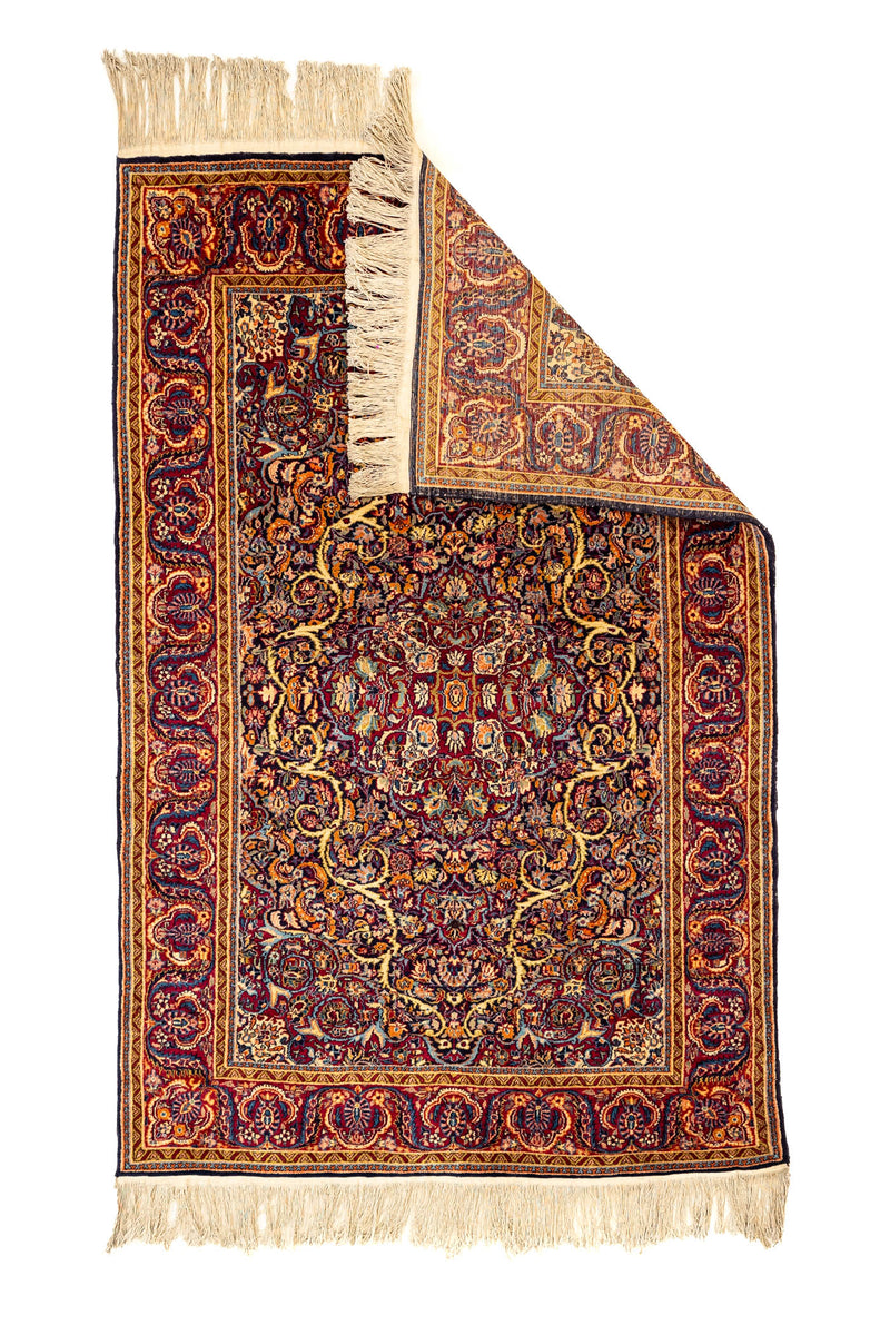 Alfombra con esquina doblada mostrando el reverso tejido y los detalles del borde floral en rojo, azul y dorado.