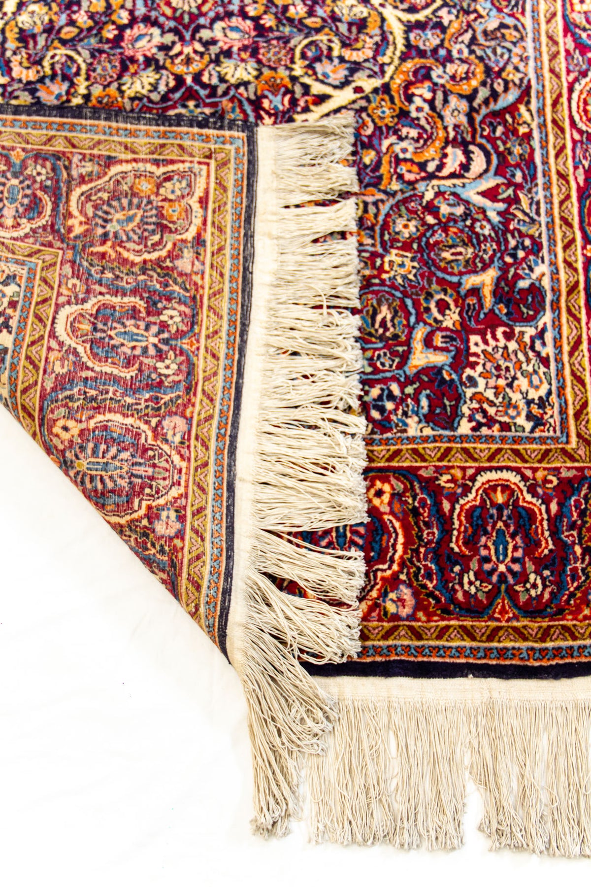 Detalle del reverso de una alfombra con flecos y diseño artesanal floral en tonos burdeos, azul y mostaza.