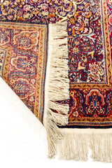 Detalle del reverso de una alfombra con flecos y diseño artesanal floral en tonos burdeos, azul y mostaza.