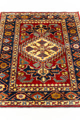 Vista inclinada de la alfombra que destaca el medallón central beige sobre el fondo rojo y los patrones en azul, dorado y verde oliva.