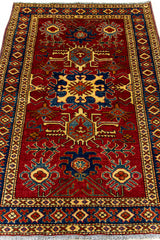 Detalle completo de la alfombra extendida, con motivos simétricos en azul, beige, dorado y verde sobre un fondo rojo intenso.