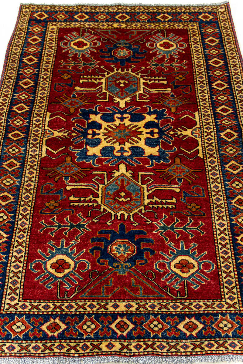 Detalle completo de la alfombra extendida, con motivos simétricos en azul, beige, dorado y verde sobre un fondo rojo intenso.